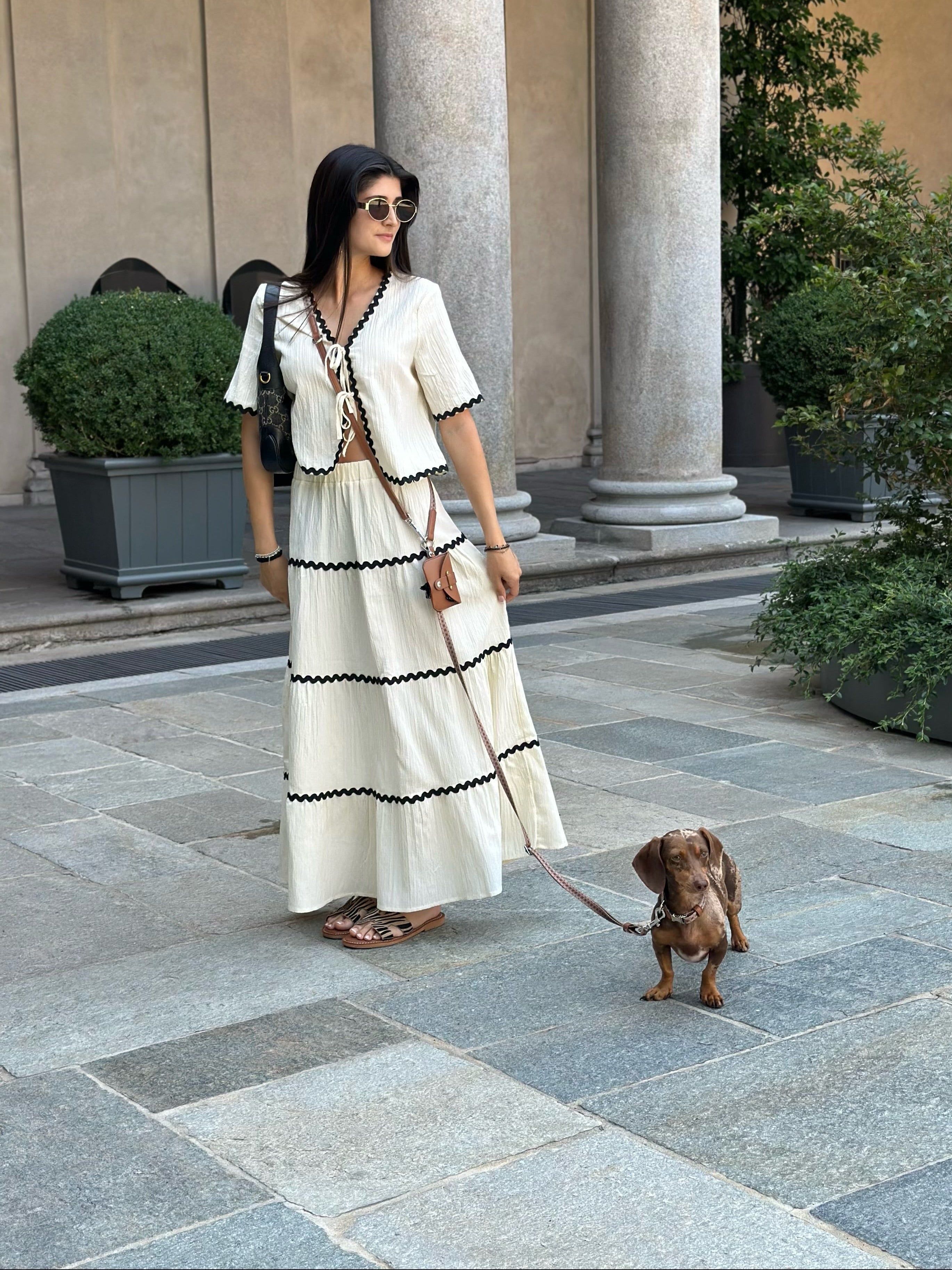 Woman in a white summer outfit walking a small dog on a multifunction leash in city.